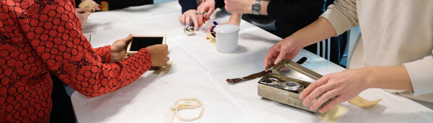 A group of MA Art Gallery and Museum Studies examine, handle and photograph museum objects and instruments laid out on a cloth-covered table.